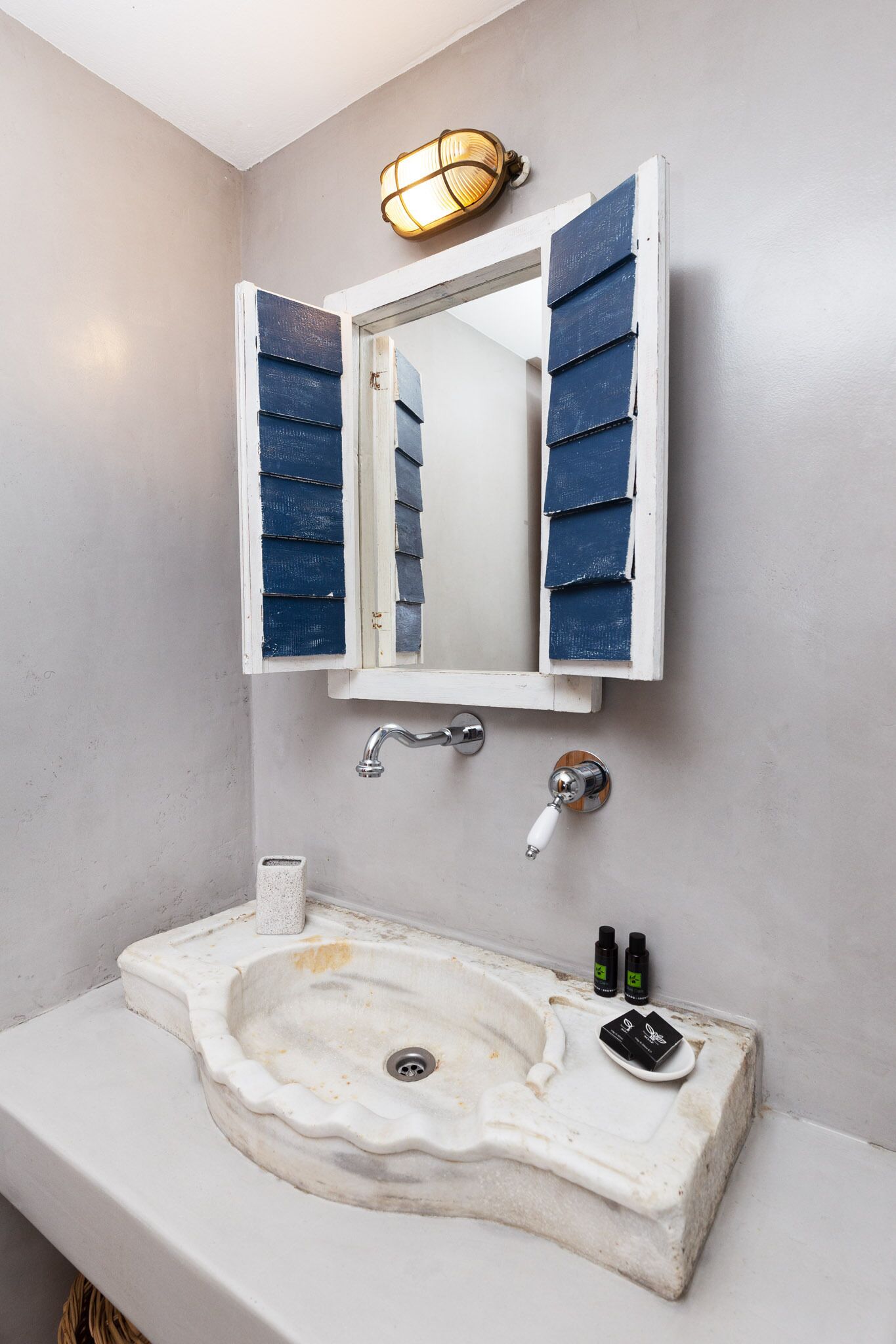 Un baño con lavamanos blanco, un espejo con persianas azules y una lámpara fijada a la pared.