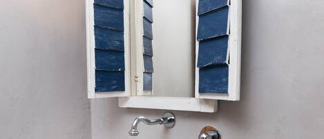 Un baño con lavamanos blanco, un espejo con persianas azules y una lámpara fijada a la pared.