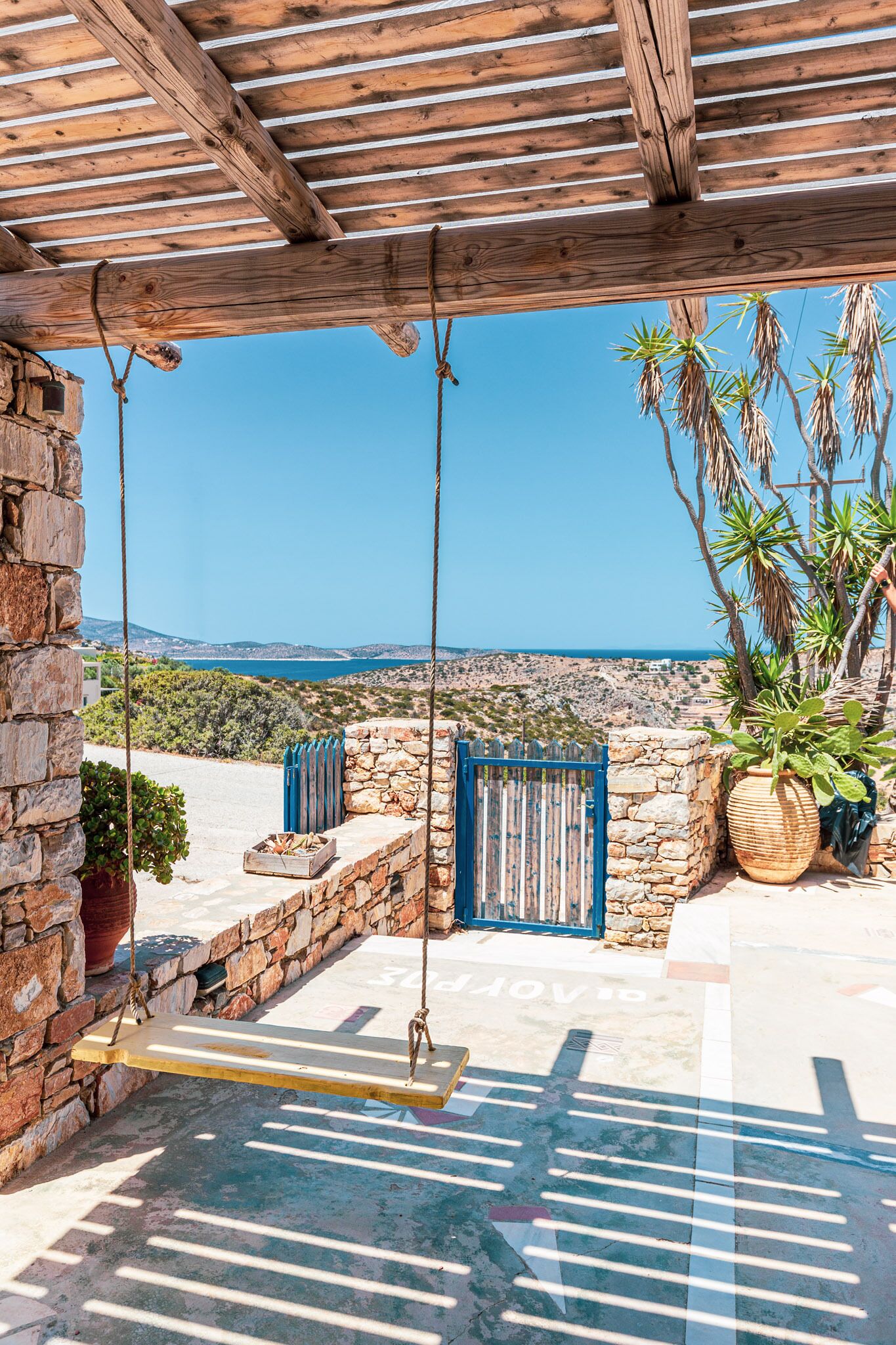 Une pergola en bois avec une balançoire, un mur de pierre, un portail bleu et une vue sur la mer et les collines.