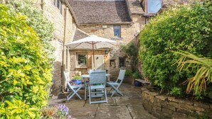 A stone building with a tiled roof, a small table with two chairs, and a potted plant. - Bull Cottage (Burford)