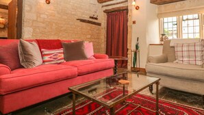 A cozy living room with a red sofa, a glass coffee table, and a beige sofa. The room features exposed wooden beams, a stone wall, and a large window with a view. - Bull Cottage (Burford)