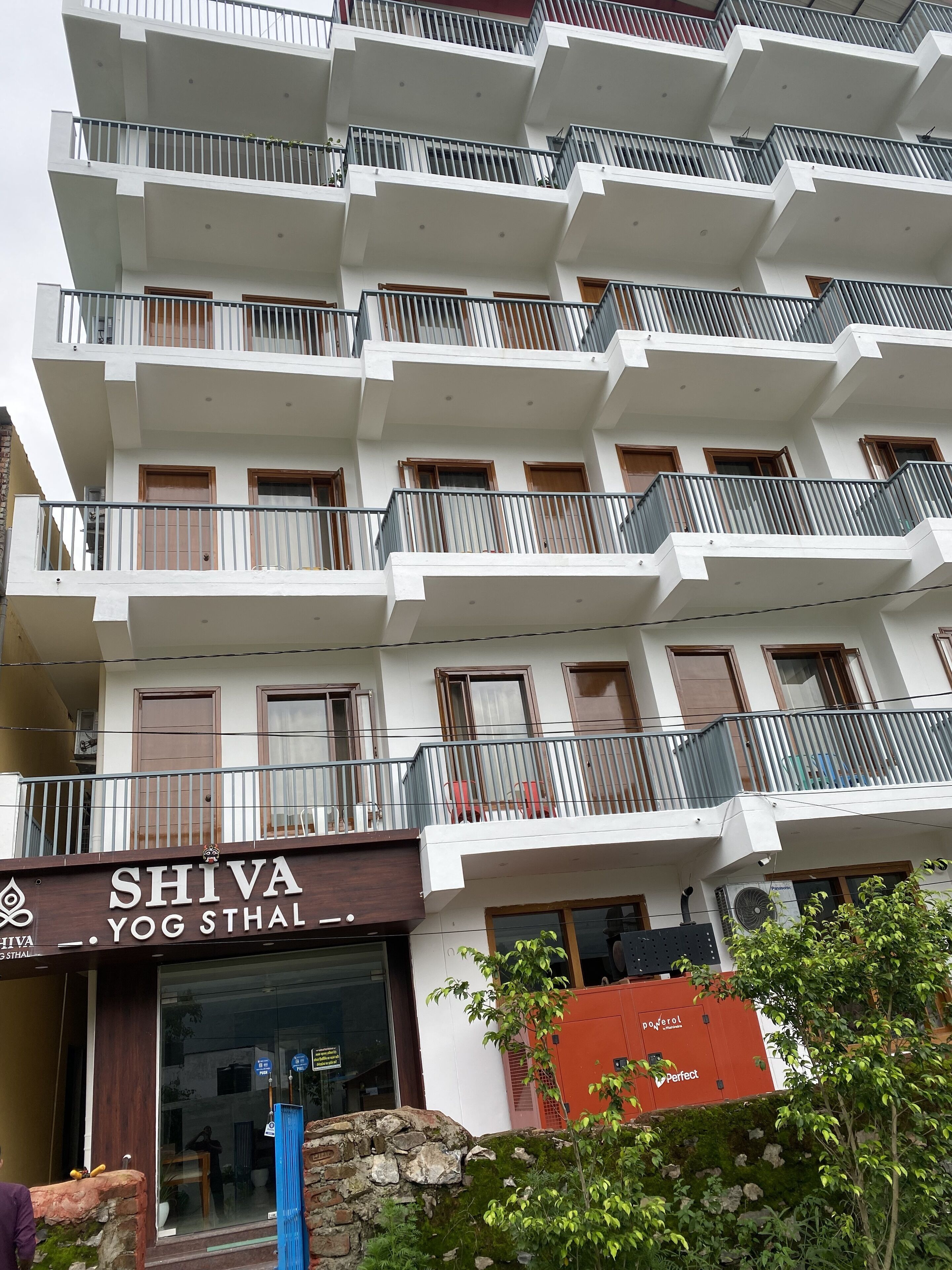 Un immeuble à plusieurs étages doté de balcons, un magasin appelé « SHIVA YOG STHAL » situé au rez-de-chaussée, et une prise de courant visible sur la façade.