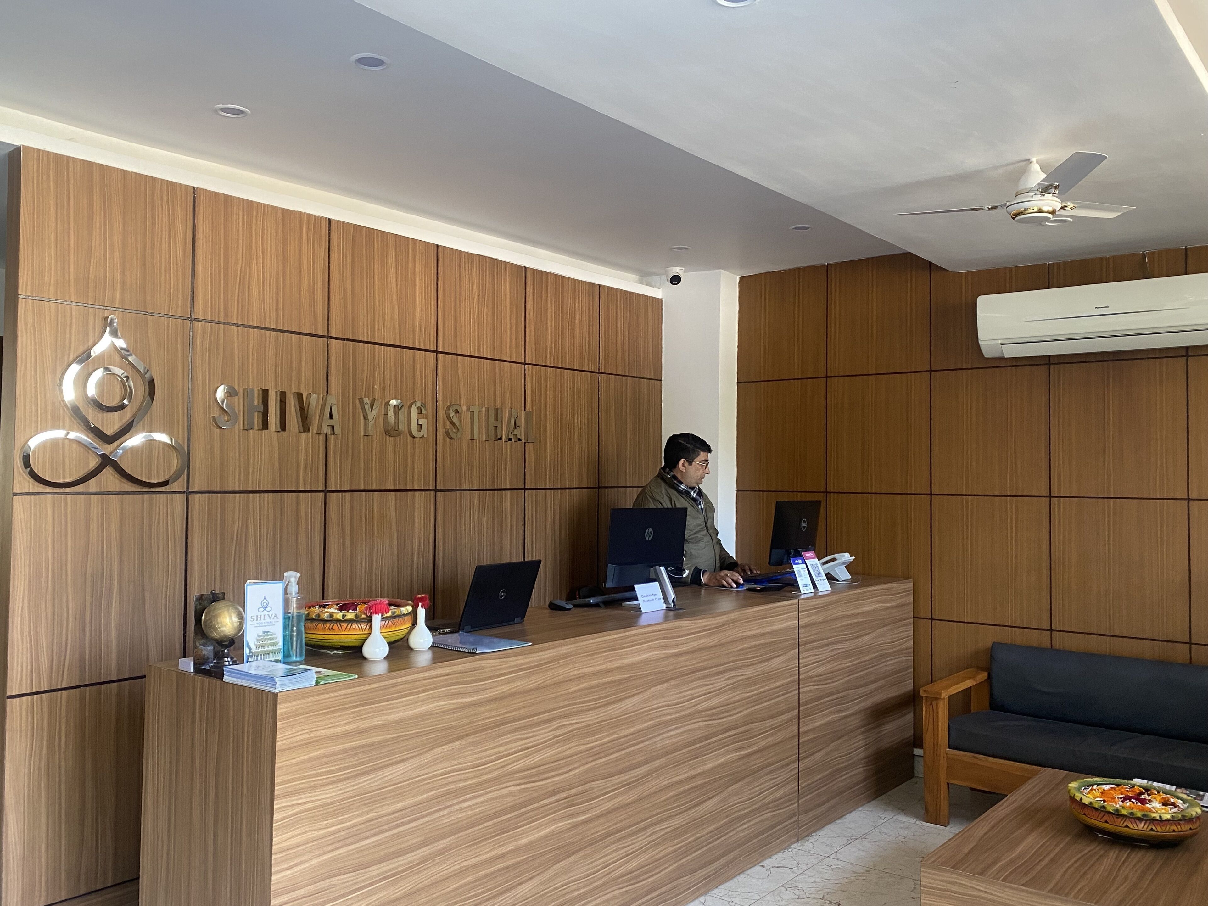 A reception desk with a wooden finish, a nameplate reading "SHIVA YOG STHAL", two laptops, a water dispenser, and a small seating area with a sofa and a table.