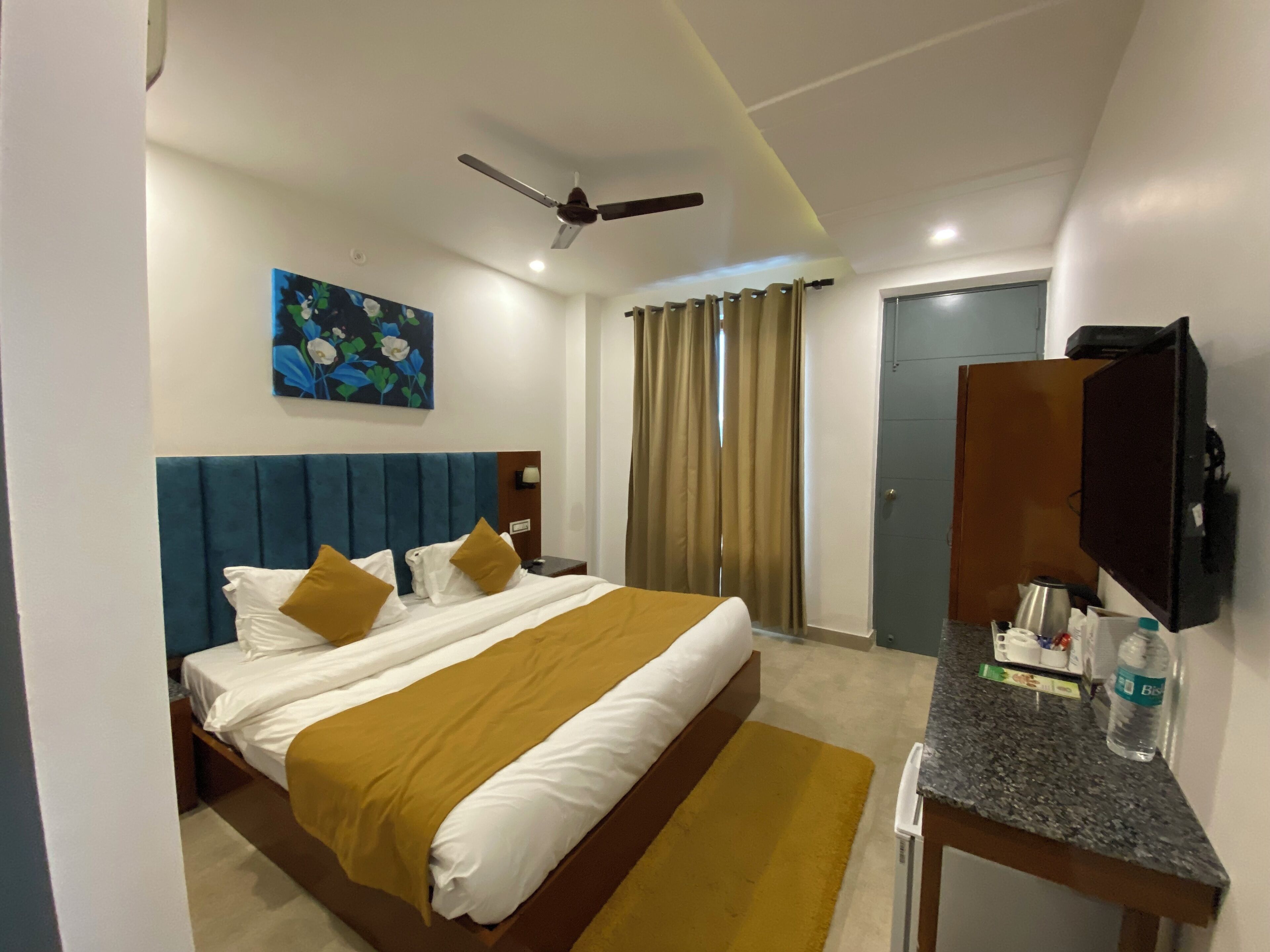 Habitación de hotel con cama, escritorio, televisor, ventilador de techo, un cuadro y una puerta.