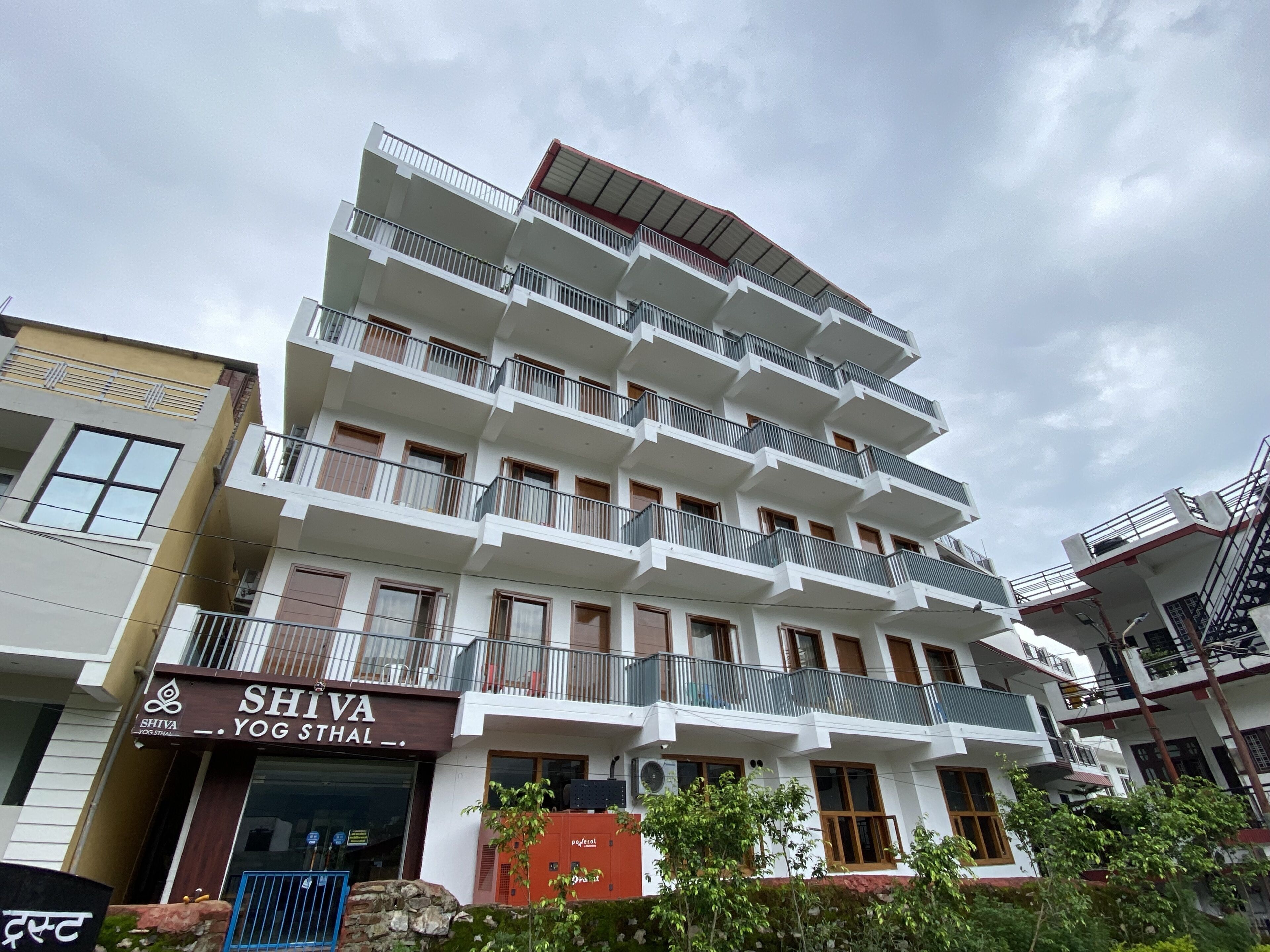 Un immeuble à plusieurs étages doté de balcons, un commerce situé au rez-de-chaussée appelé « SHIVA YOG STHAL », et un coffret électrique rouge.