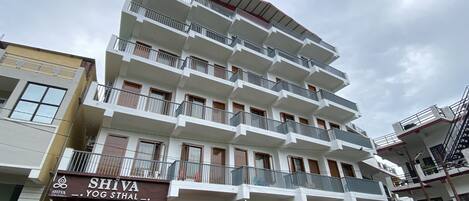 Un immeuble à plusieurs étages doté de balcons, un commerce situé au rez-de-chaussée appelé « SHIVA YOG STHAL », et un coffret électrique rouge.