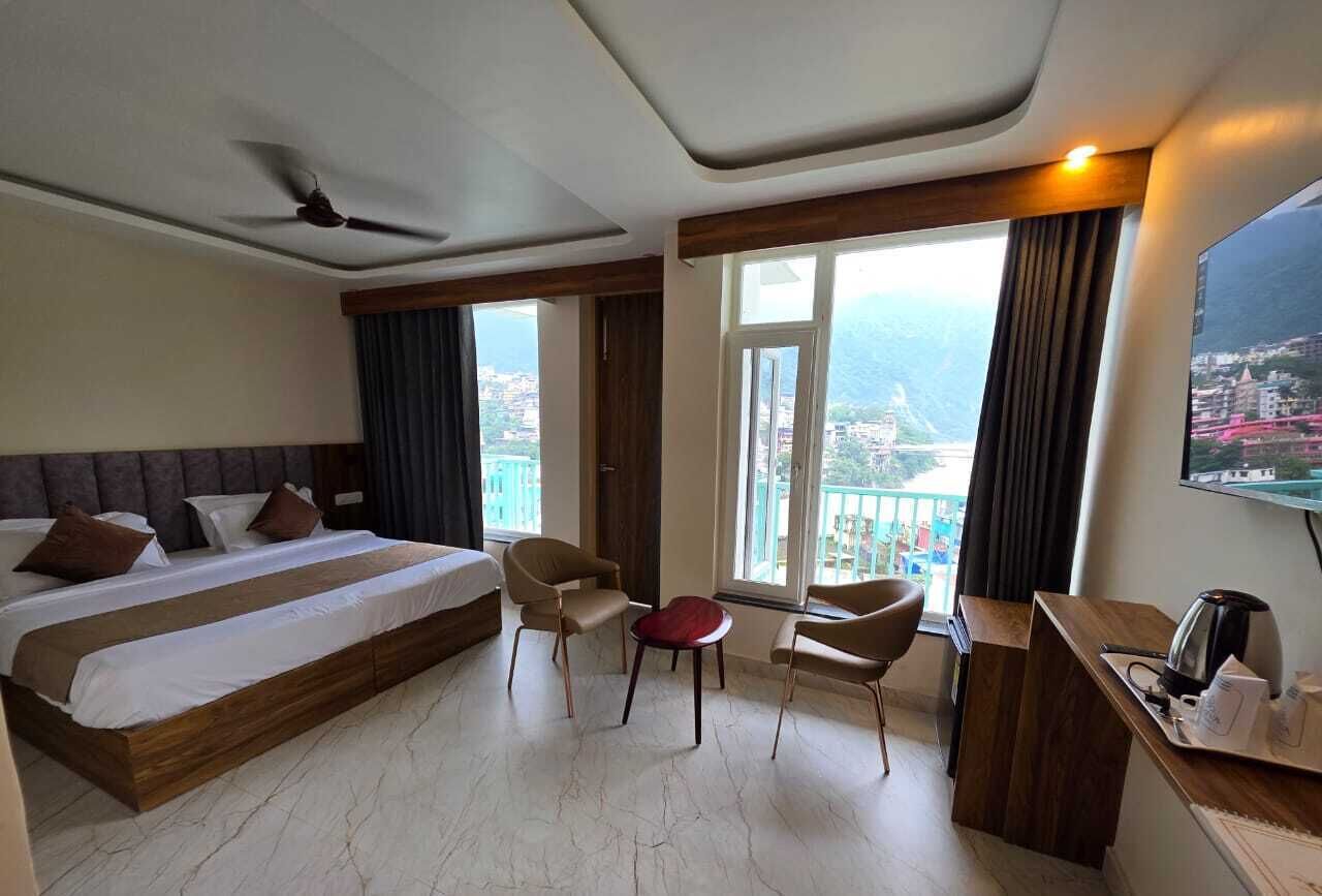 A hotel room with a bed, two chairs, a small table, a desk with a coffee maker, and a large window with a view of a city and mountains.