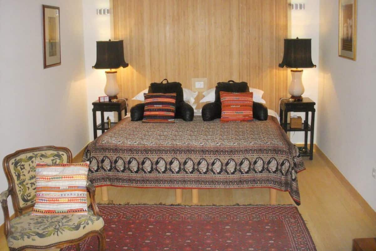 Un dormitorio con una cama grande, dos mesitas de noche con lámparas, una silla con cojín estampado y un cabecero de madera.