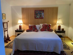 Un lit bien fait, agrémenté de coussins à motifs floraux, de deux lampes de chevet, d’une œuvre d’art encadrée au-dessus du lit et d’une tête de lit en bois.