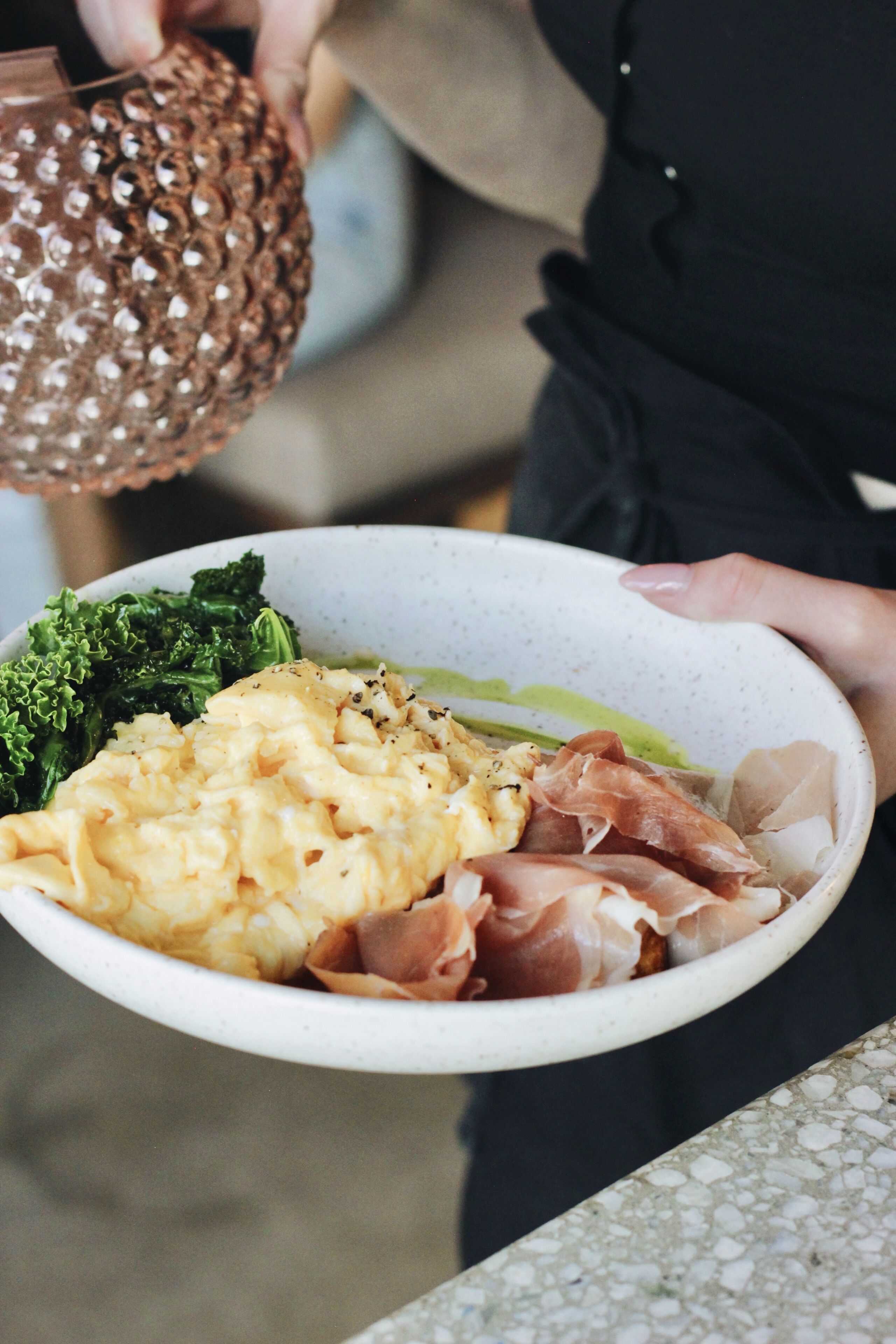 A person holding a bowl with food items including broccoli, scrambled eggs, and prosciutto.