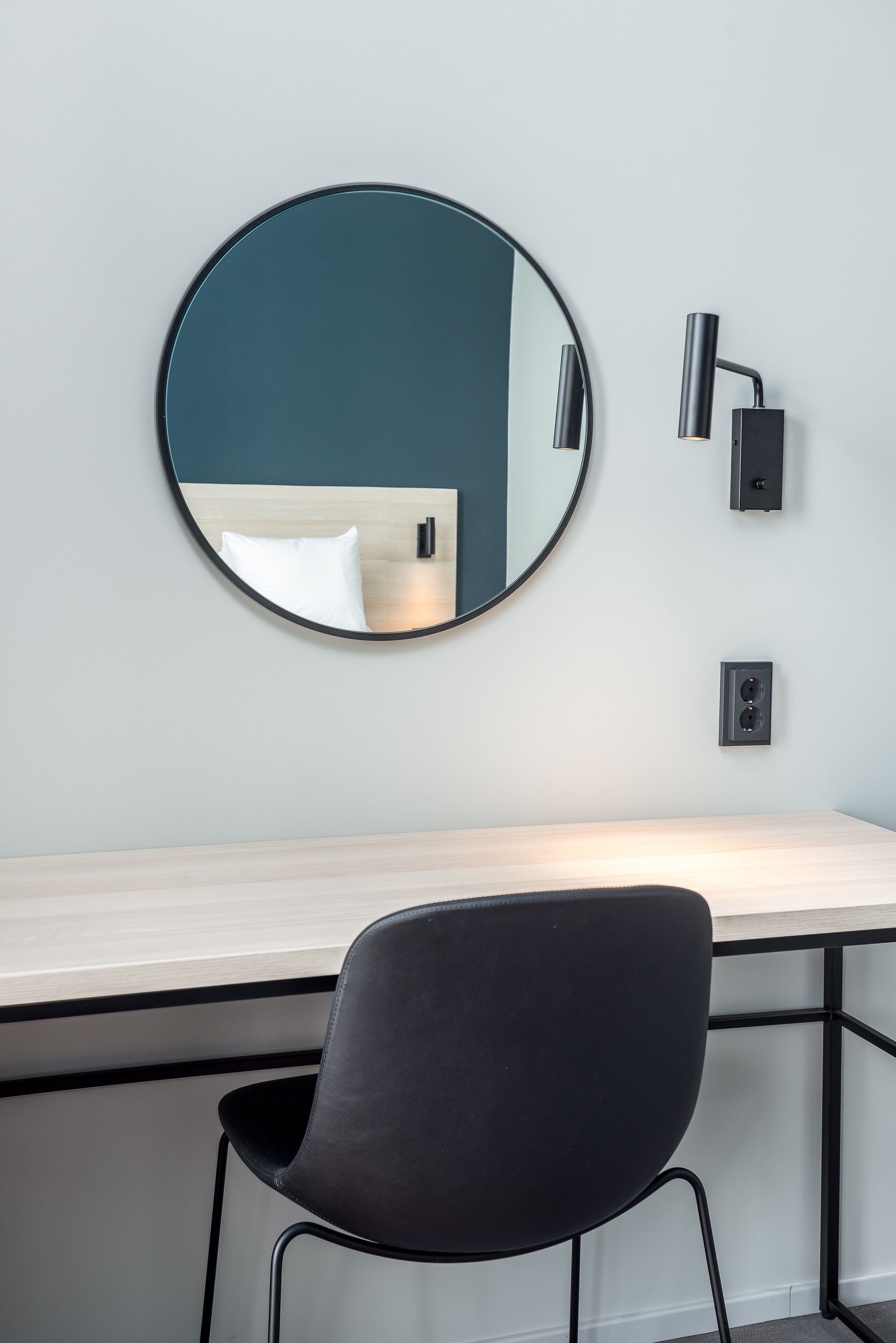 A modern room with a round mirror on the wall, a desk with a chair, and a wall-mounted lamp.