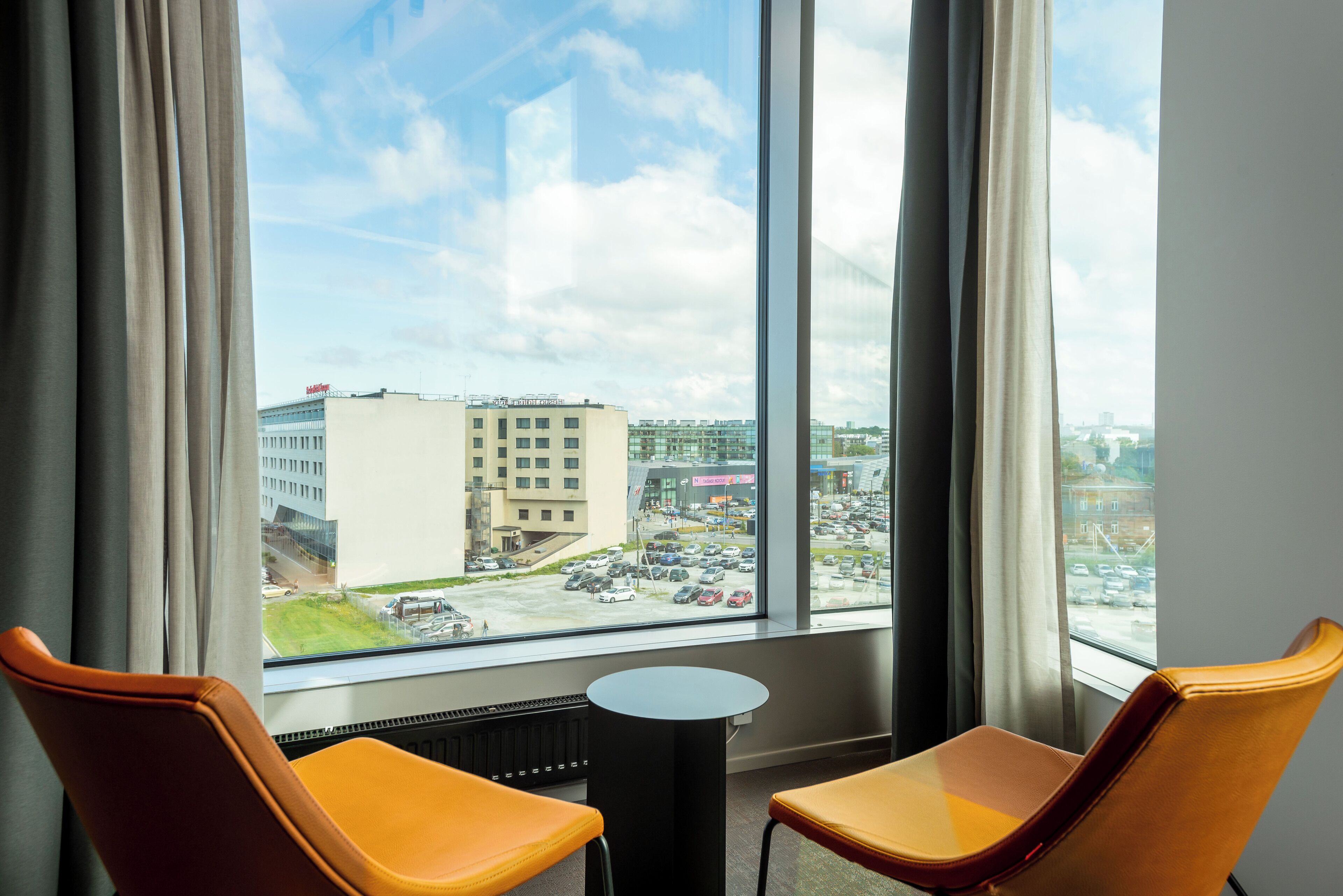 A hotel room with two orange chairs, a small round table, and a large window offering a view of a parking lot and buildings.