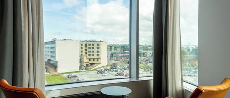 A hotel room with two orange chairs, a small round table, and a large window offering a view of a parking lot and buildings.