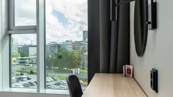 A hotel room with a large window, a wooden desk, a black chair, and a wall-mounted light fixture.