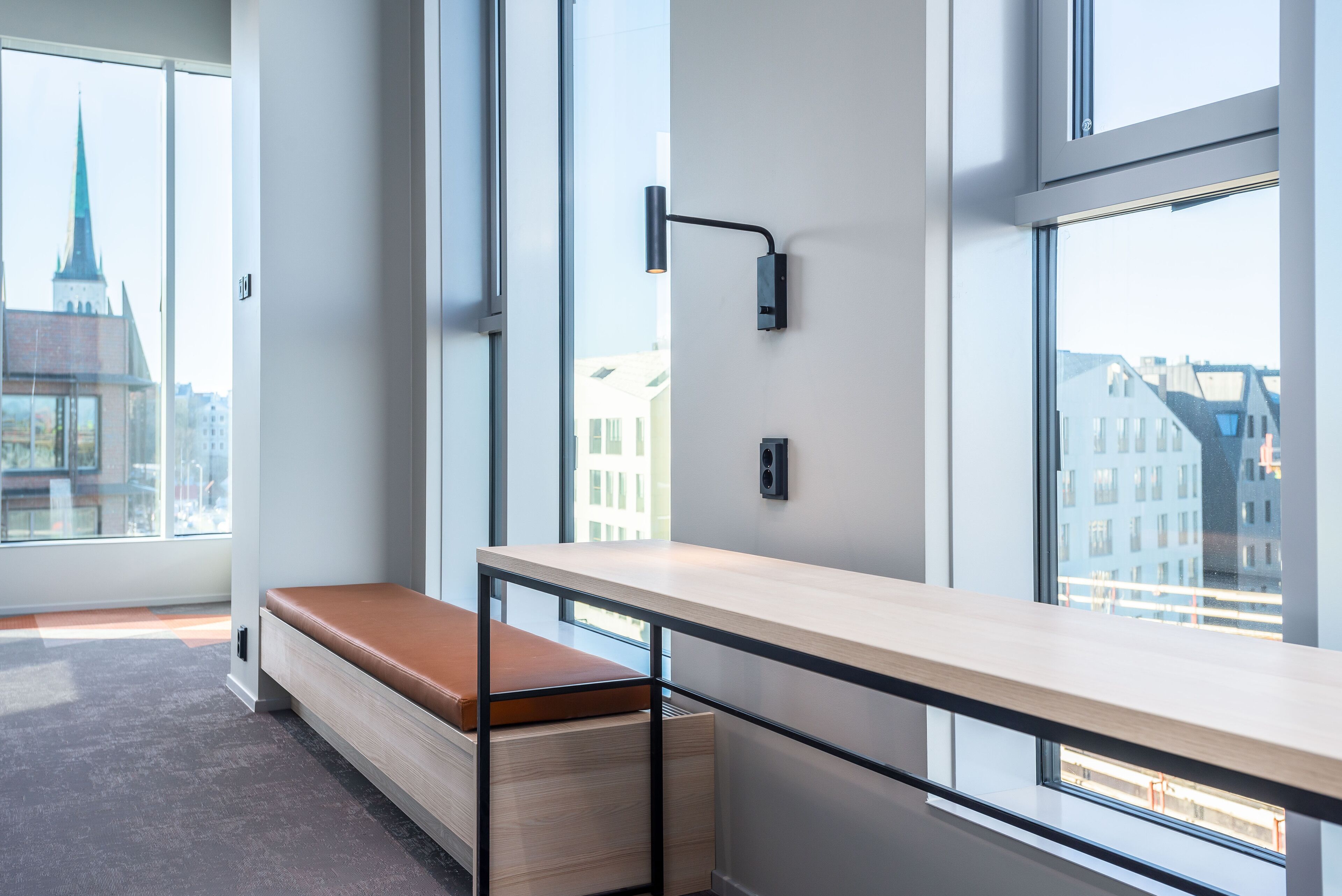 A modern room with a wooden bench, a desk with a light fixture, large windows, and a view of buildings.