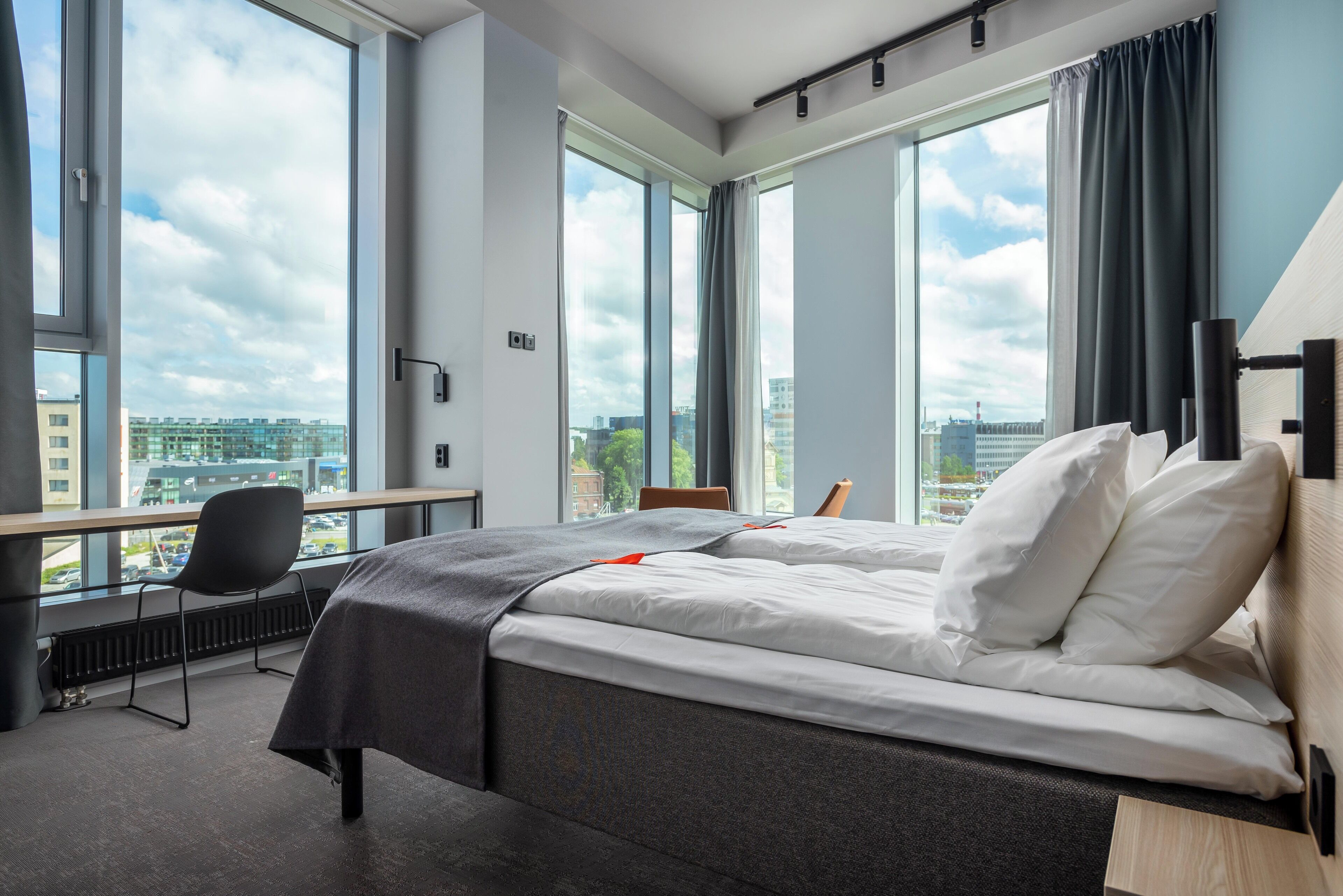 A hotel room with a bed, a desk, and a chair, large windows with a city view, and a view of buildings outside.
