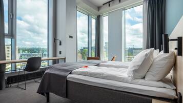 A hotel room with a bed, a desk, and a chair, large windows with a city view, and a view of buildings outside.