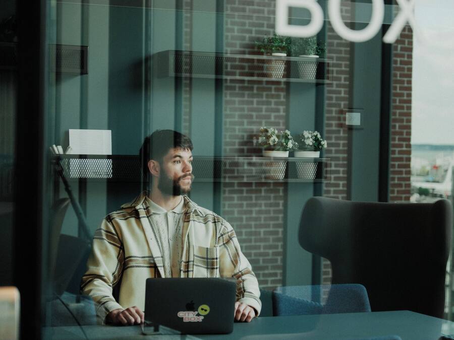 A person sitting at a table with a laptop, in a room with a glass wall, a brick wall, and shelves with plants.