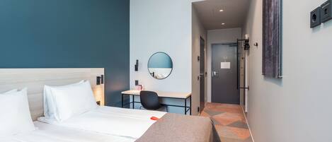 Quarto de hotel com cama, escrivaninha, cadeira, espelho e porta que dá para outro cômodo.