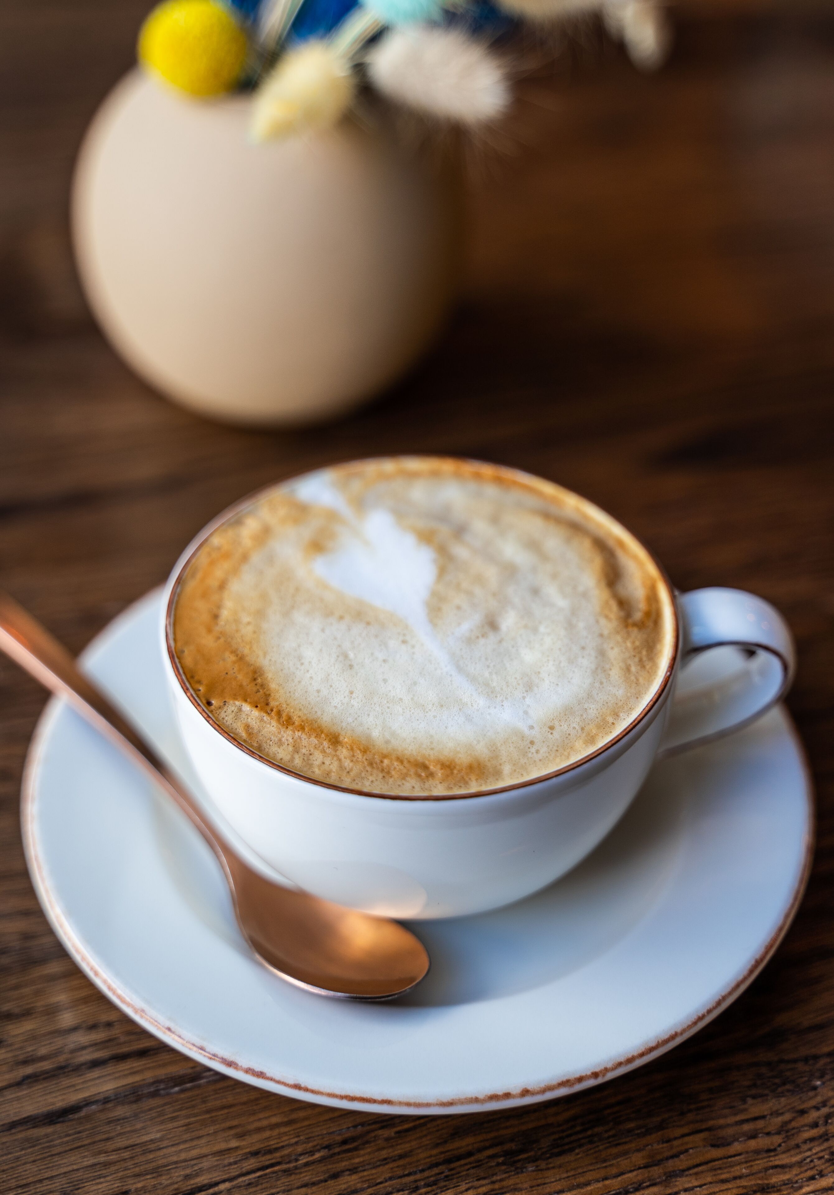 A cup of coffee with latte art on a saucer, a spoon, and a blurred vase in the background.