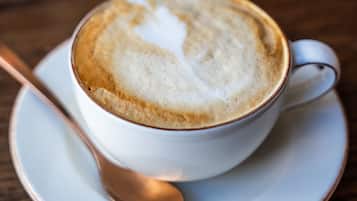A cup of coffee with latte art on a saucer, a spoon, and a blurred vase in the background.