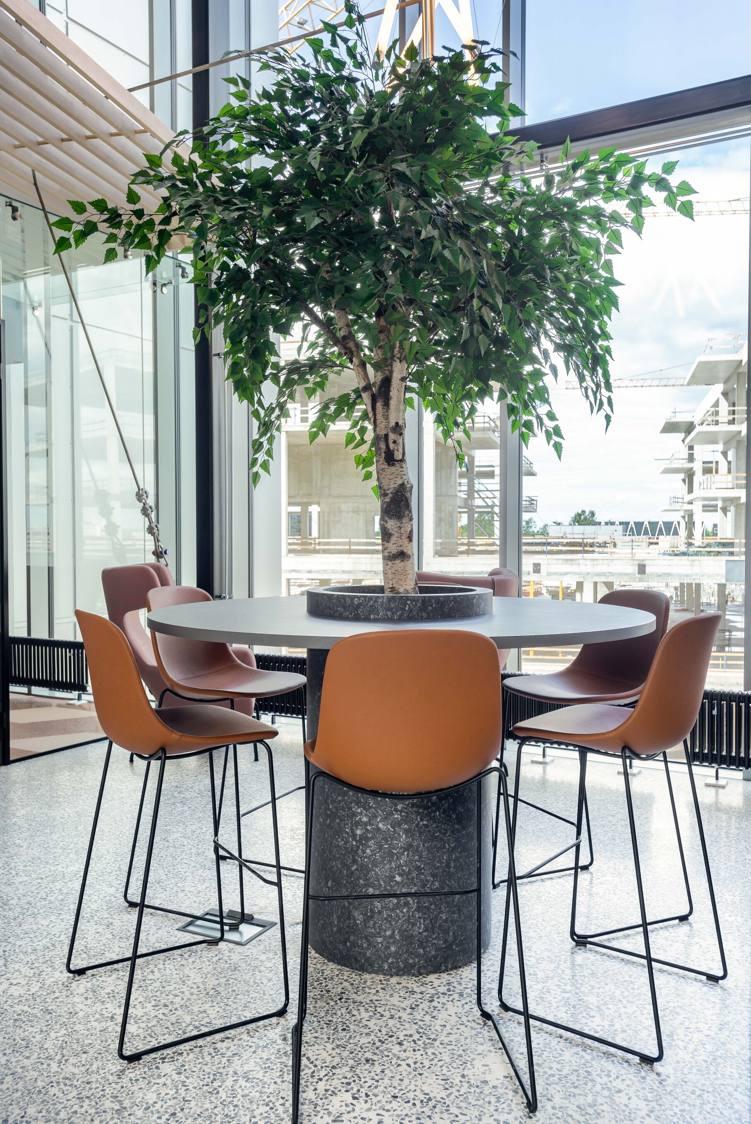 A modern indoor setting with a round table surrounded by orange chairs, a central potted tree, and large windows offering a view of buildings and construction.