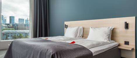 Una habitación de hotel moderna con una cama grande, una mesita de noche y vistas de la ciudad a través de un amplio ventanal.