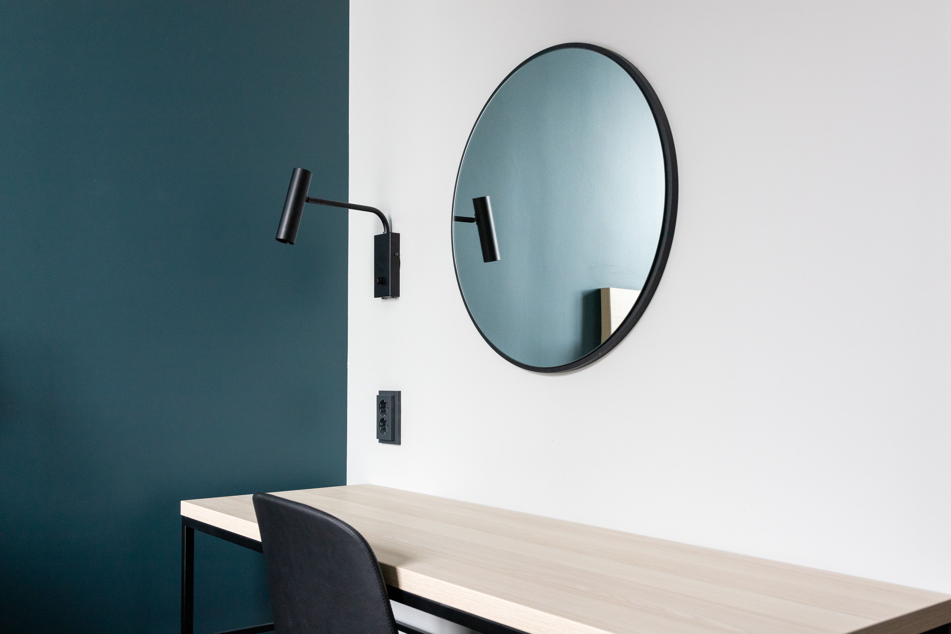 A modern room with a wooden desk, a black chair, a round mirror, and a wall-mounted light fixture.