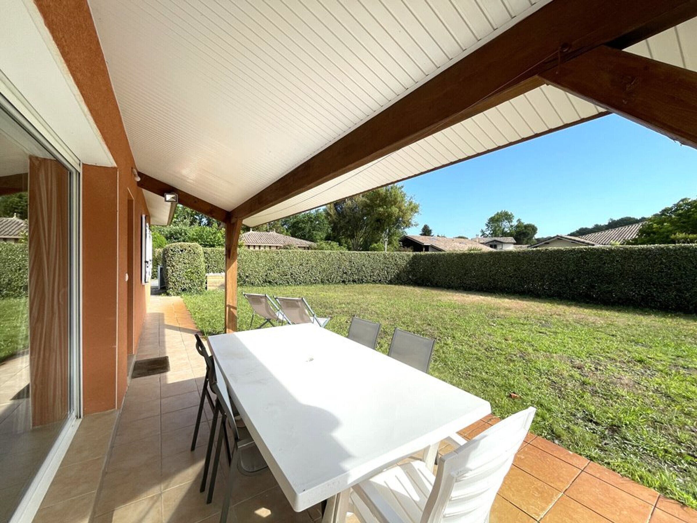 Une terrasse extérieure avec une table et des chaises blanches, un espace couvert doté d’un sol carrelé, et une vue sur une pelouse bien entretenue et des bâtiments situés au loin.