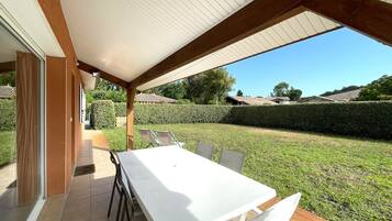 Une terrasse extérieure avec une table et des chaises blanches, un espace couvert doté d’un sol carrelé, et une vue sur une pelouse bien entretenue et des bâtiments situés au loin.