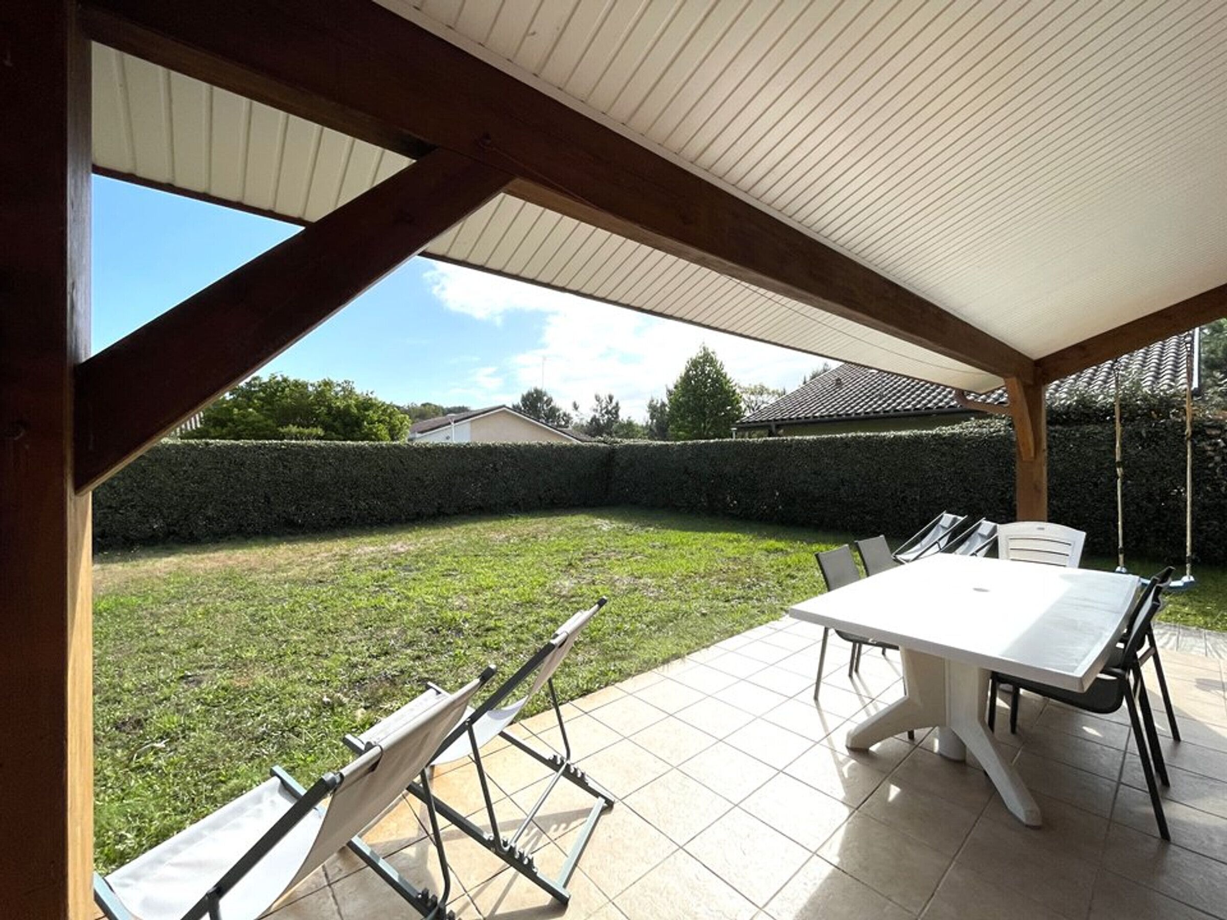 Uma área de varanda coberta com mesa e cadeiras, vista para um gramado e um edifício ao fundo.