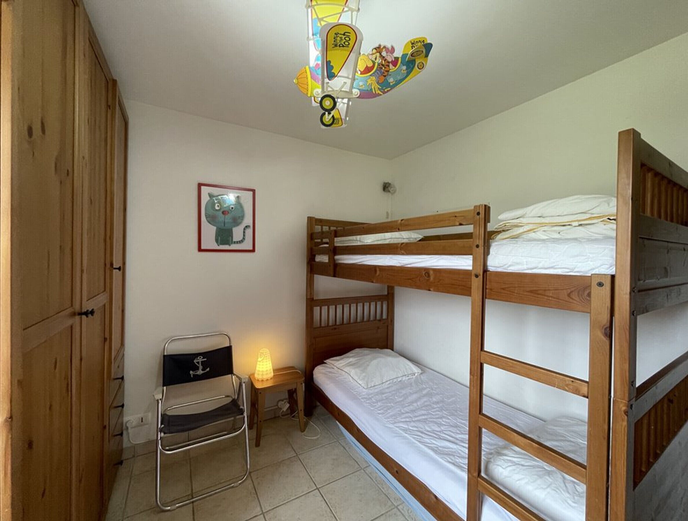 Een slaapkamer met een stapelbed, een houten kledingkast, een klein bureau met stoel en een wandlamp.