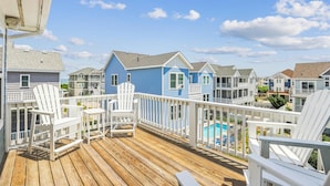 Eine Holzterrasse mit weißen Adirondack-Stühlen, einem Pool und einem blauen Haus in einer Wohngegend an der Küste.