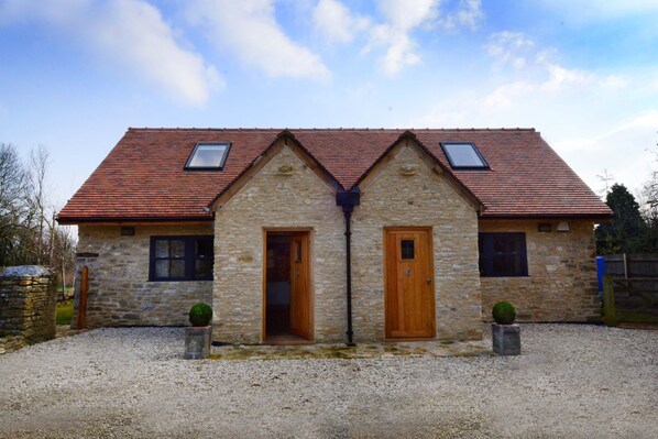 A stone building with a red-tiled roof, two windows, and a gravel driveway. - Dove House Cottage 1 (Witney)