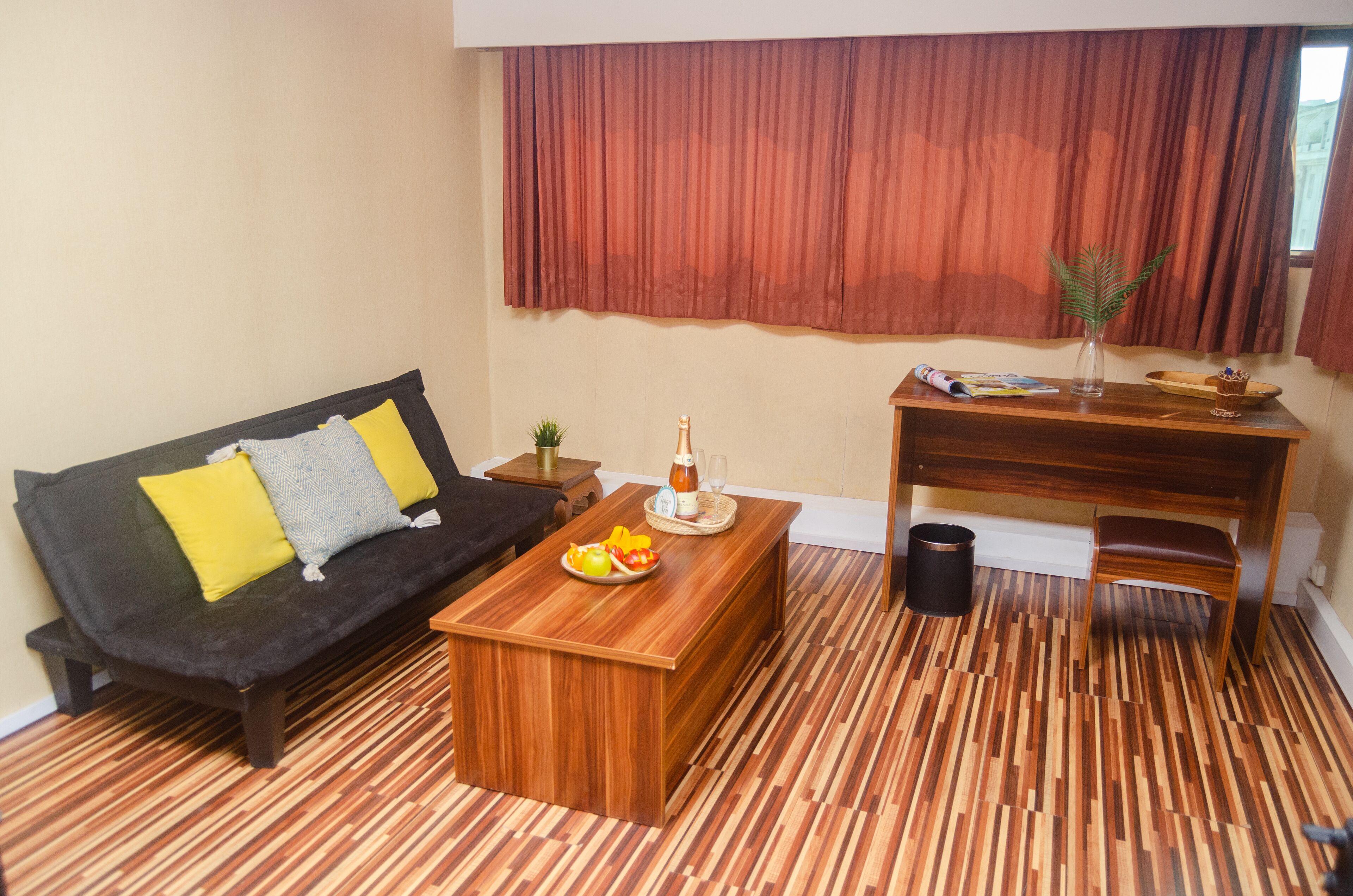 A living room with a dark sofa, wooden coffee table, and a wooden desk with a chair. A small plant is on the desk, and there is a trash can next to it.