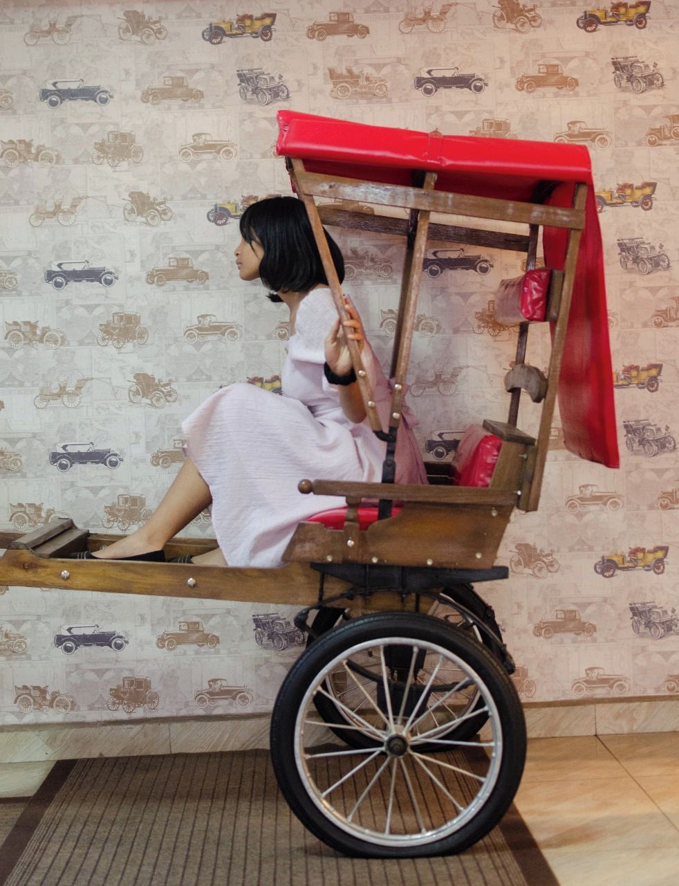 A person seated in a wooden cart with a red canopy, against a wallpapered wall with a pattern of vehicles.