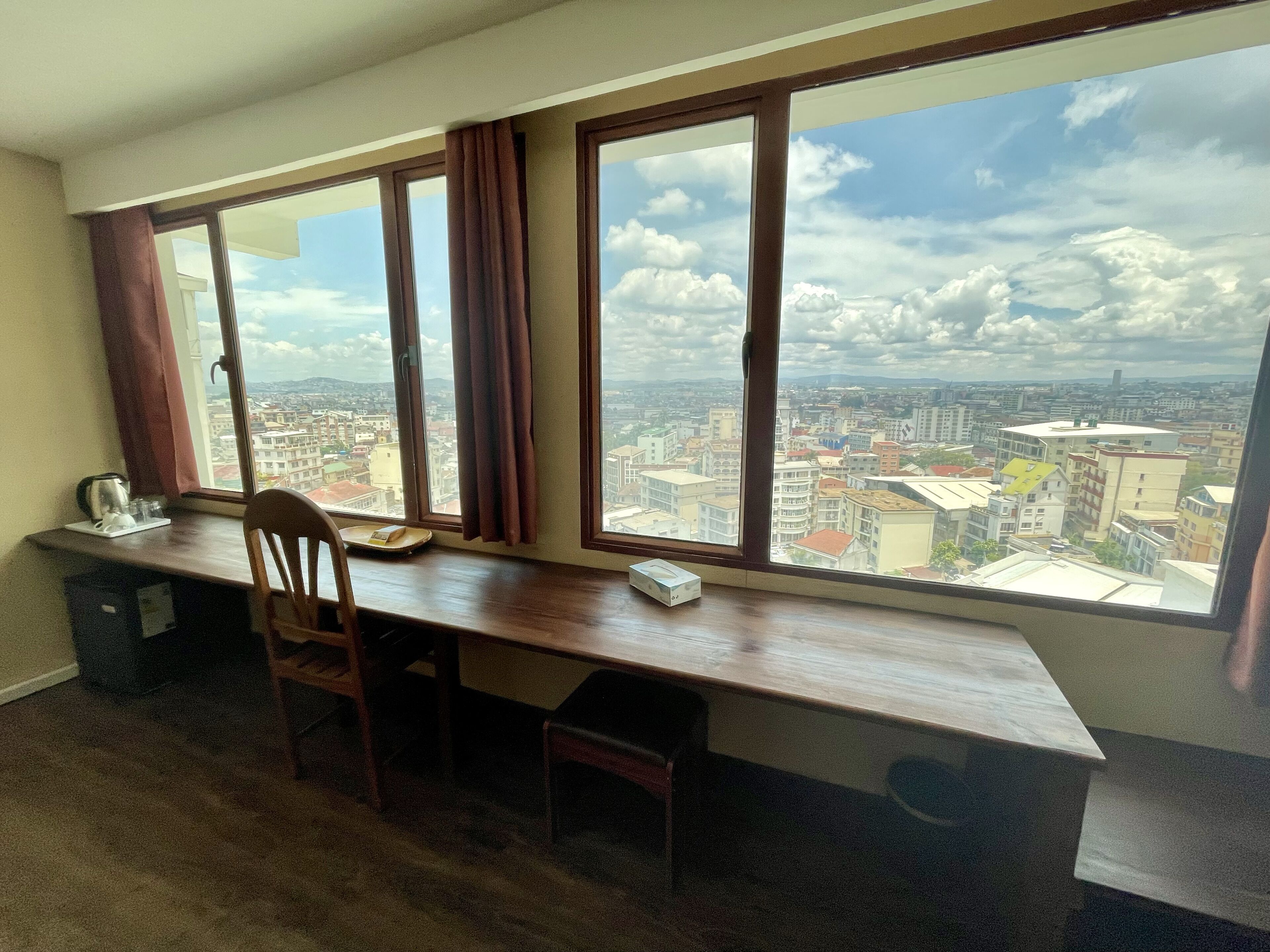 Ein Zimmer mit großem Fenster, einem Holztisch, einem Stuhl und Blick auf eine Stadtansicht.