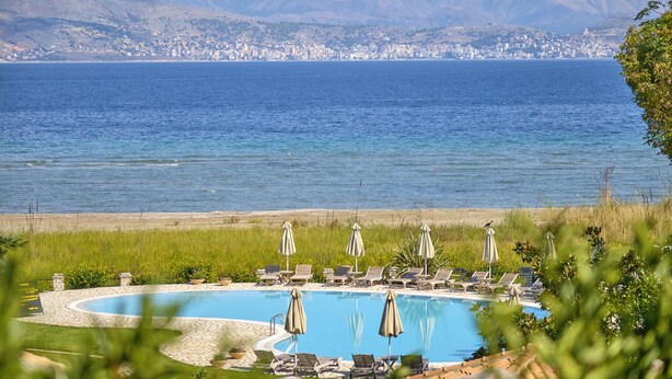 Ein Pool mit Liegestühlen und Sonnenschirmen, umgeben von einer angelegten Grünfläche mit Blick auf das Meer und eine entfernte Stadtlandschaft.