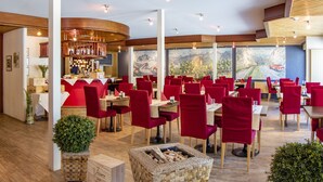 Ein Restaurant mit roten Stühlen, Holztischen und Blick auf eine Berglandschaft durch große Fenster.