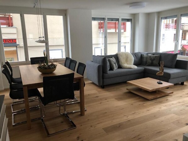A modern living room with a dining area, large windows, and a view of the outside.