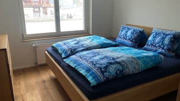 Un dormitorio con cama de madera, ropa de cama azul estampada, una ventana con vistas a edificios y un radiador.