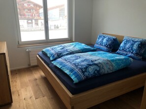 A bedroom with a wooden bed, blue patterned bedding, a window with a view of buildings, and a radiator.