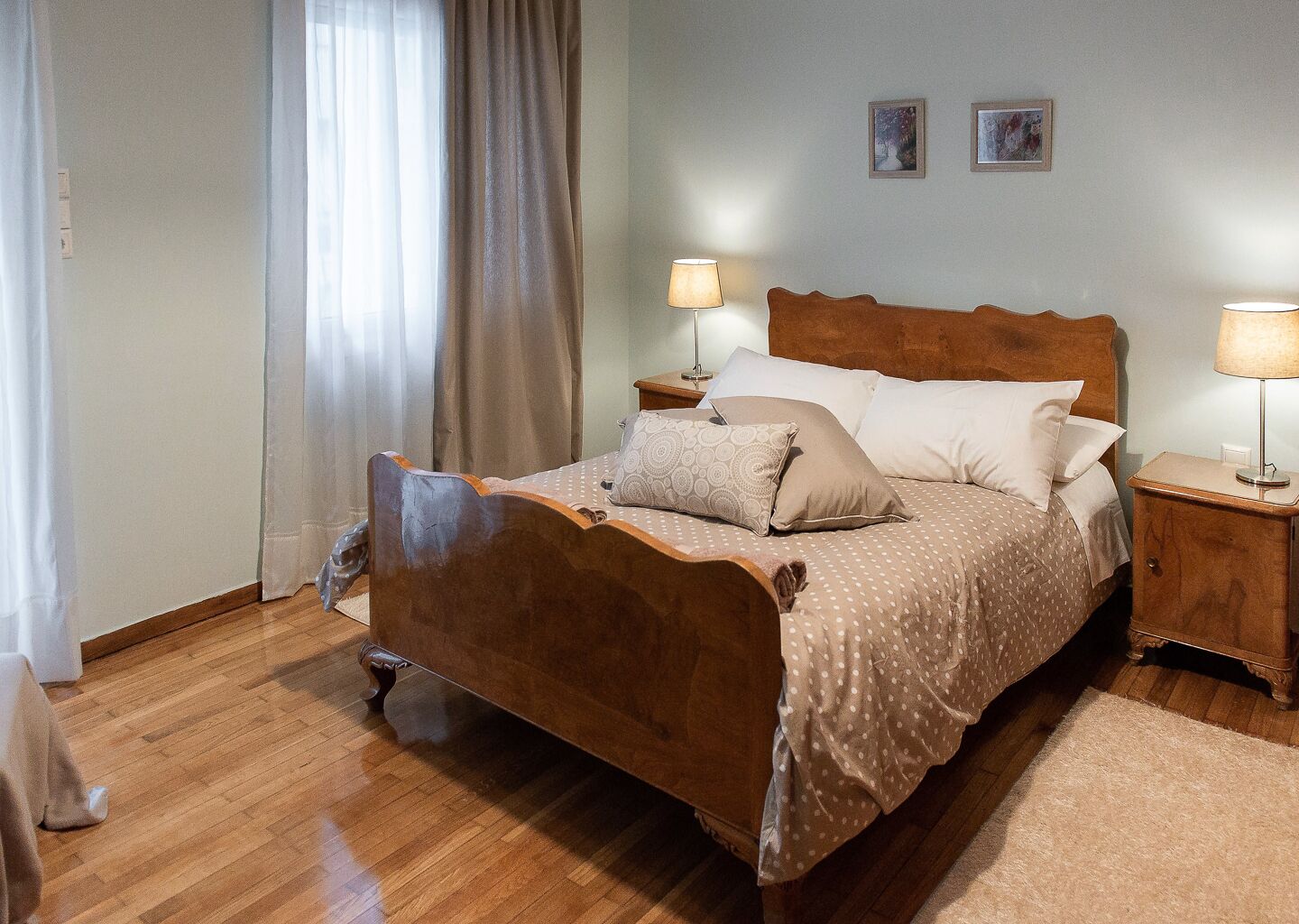 Un dormitorio con una cama de madera, dos mesitas de noche con lámparas, una ventana con cortinas y dos cuadros en la pared.