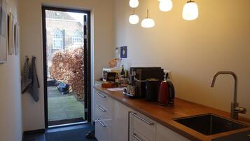 Een moderne keuken met een inbouwgootsteen, een koffieapparaat en een waterkoker op het aanrecht. Een deur leidt naar een tuin met een gebouw van baksteen op de achtergrond.