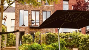 Een bakstenen gebouw met een balkon, omgeven door weelderige begroeiing en een terras met stoelen en een parasol.