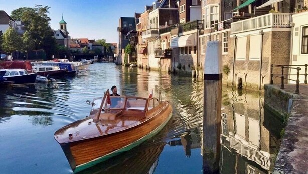 Een smalle gracht met een houten bootje, gebouwen aan de rechterkant en een kerktoren in de verte.