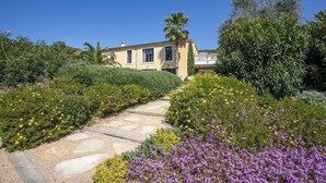 A Mediterranean-style villa with a stone pathway leading to the entrance, surrounded by lush greenery and flowering plants. - Luxurious finca with large pool in a unique location (Andratx)