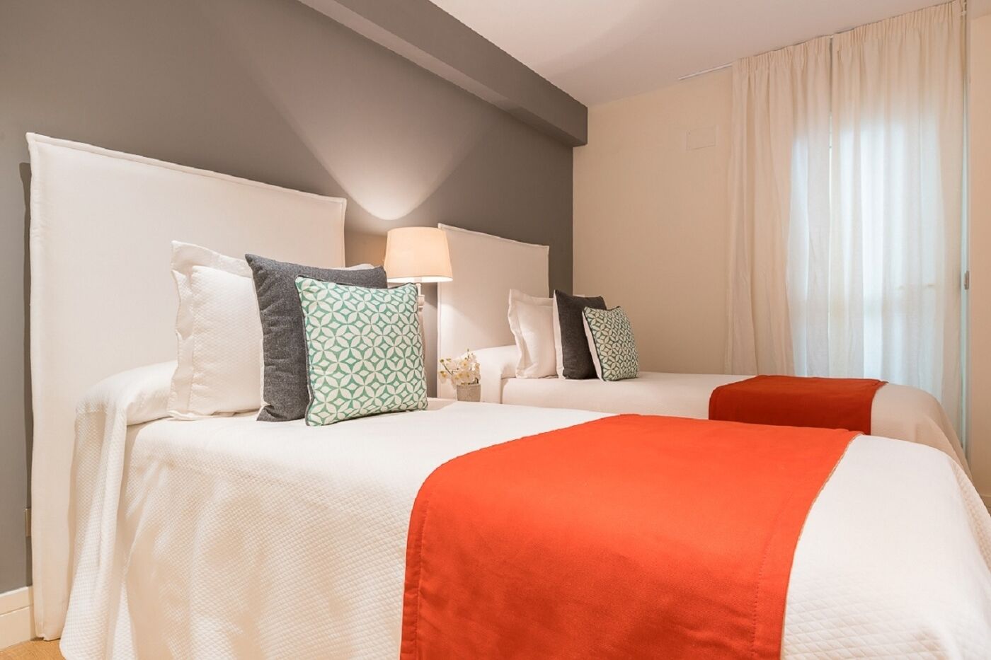 Ein Hotelzimmer mit zwei Betten, jeweils mit weißer Bettwäsche und orangefarbenen Zudecken. Mehrere Kissen, darunter gemusterte. Eine Lampe steht auf dem Nachttisch, und das Zimmer hat eine neutrale Farbgestaltung mit beige Wänden und einem Fenster mit durchsichtigen Vorhängen.