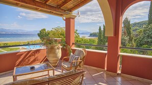 A balcony with wicker furniture, a potted plant, and a view of the sea and hills.