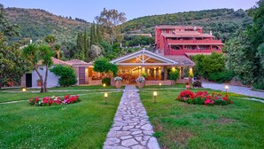 A hotel with a stone pathway leading to a covered patio area, surrounded by well-maintained gardens and a mountainous backdrop.
